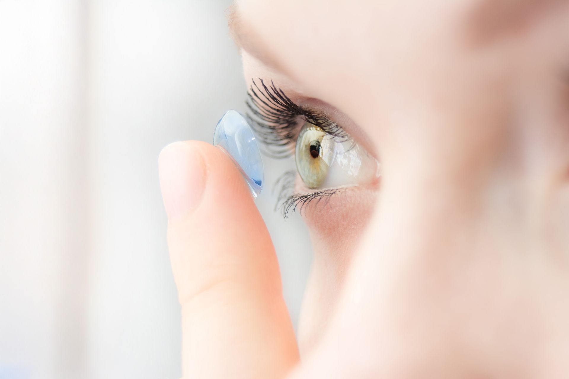 Person inserting a contact lens into their eye, close-up with the lens visible, fingers, eye with light green iris.