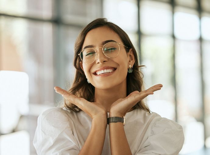 Woman with glasses smiles widely, holding her hands up near her face.