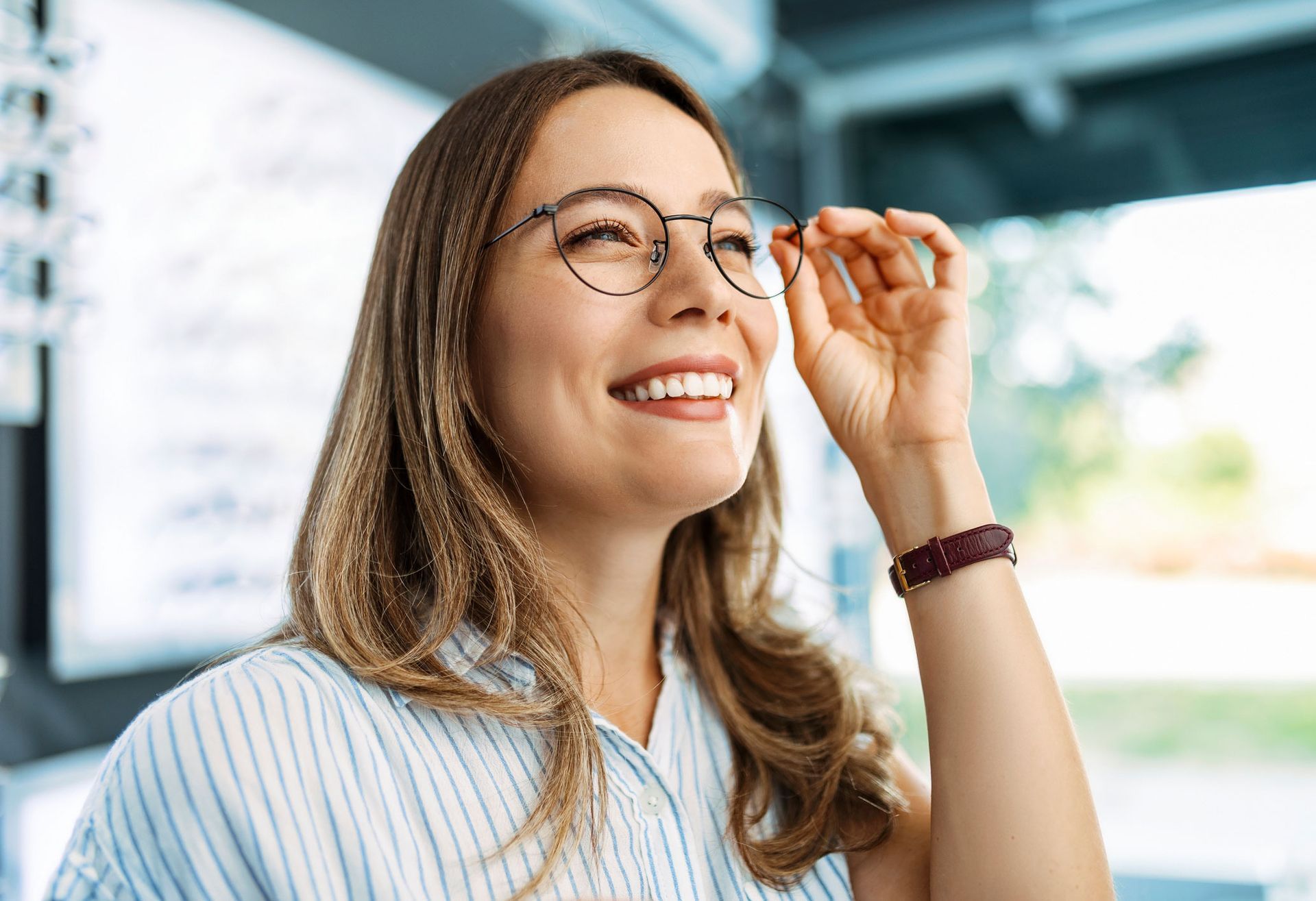 Woman smiling, adjusting round glasses, light blue striped shirt, in a store.