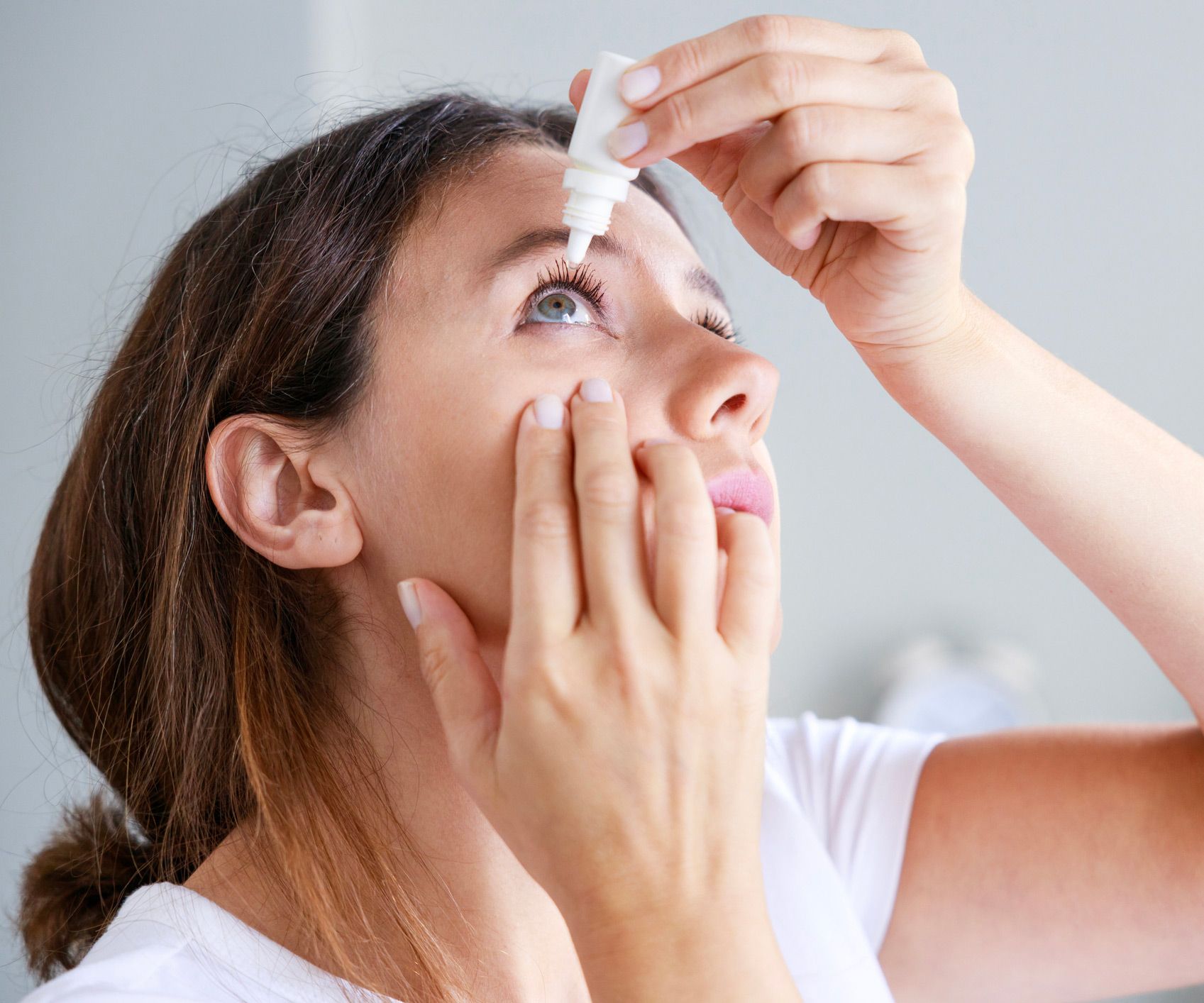 Woman applying eye drops, looking up. Indoors, pale skin, white shirt.