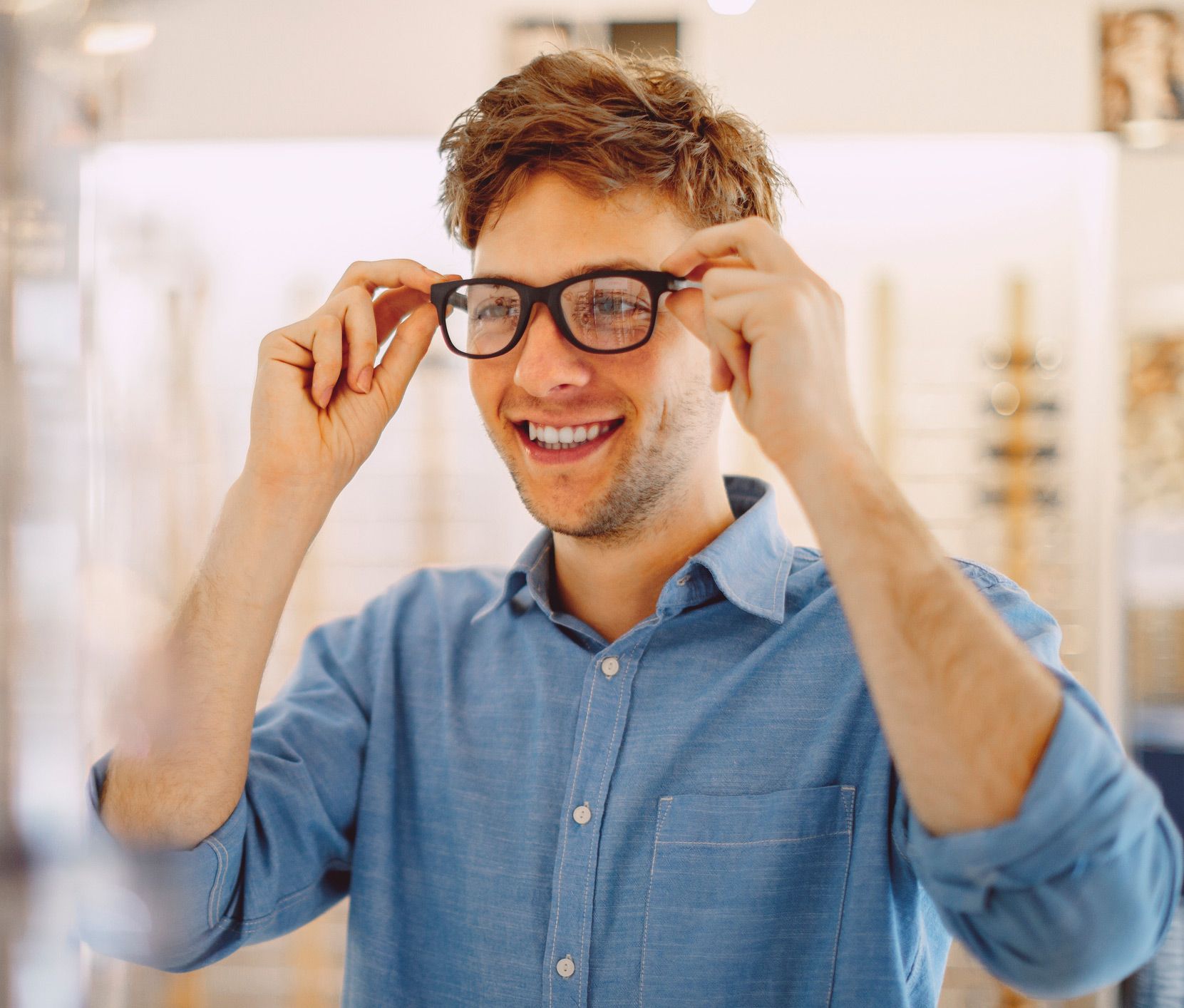 Man in blue shirt smiles, trying on black framed glasses in an optical shop.