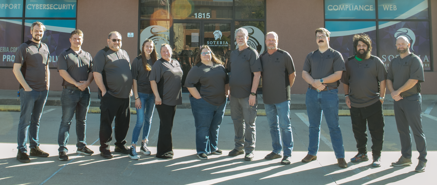 A line of several men and women in matching grey shirts with a Soteria logo on them line up in front of the Soteria office.