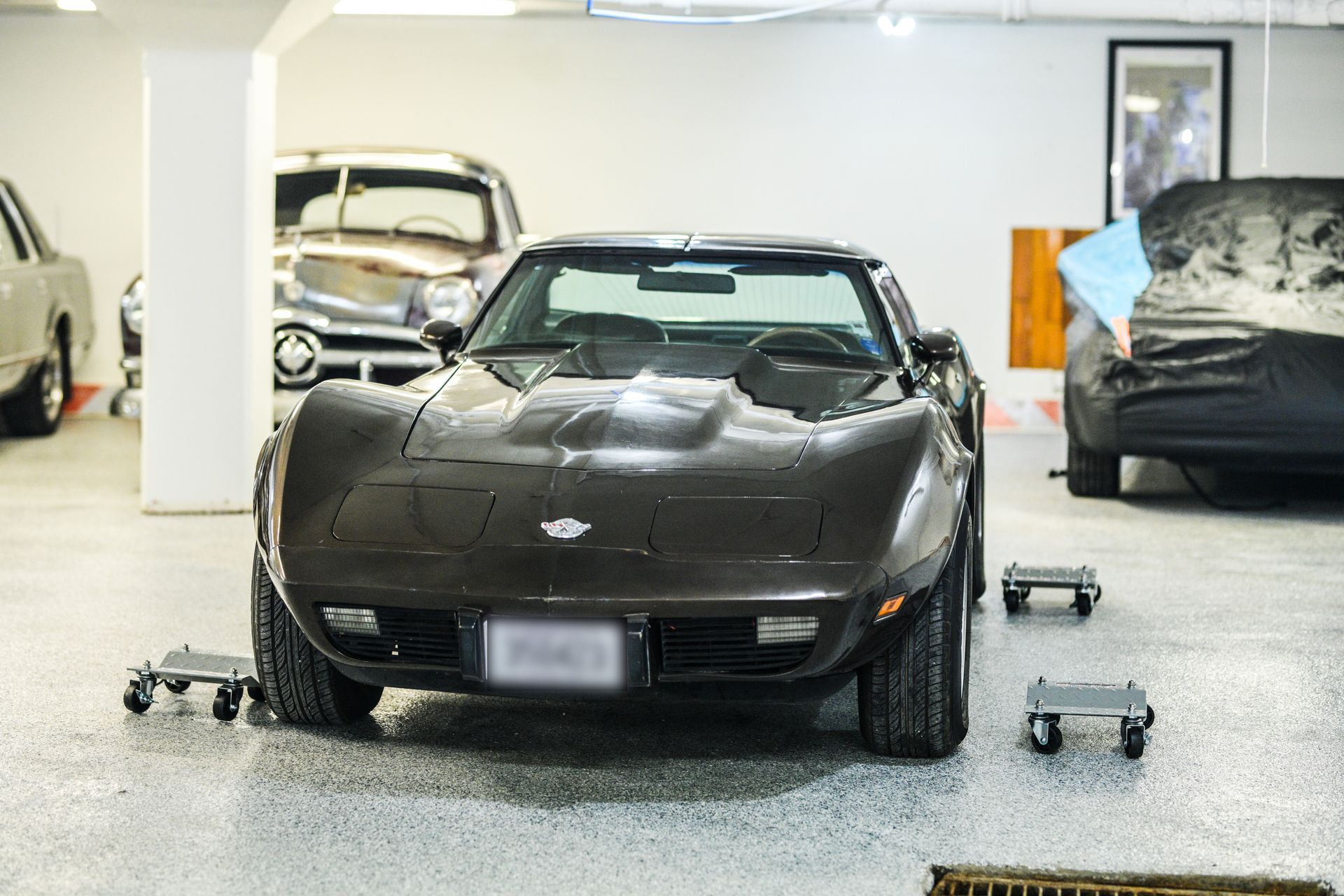 A dark brown vintage Corvette sports car parked in a garage. Two other cars are visible in the background.