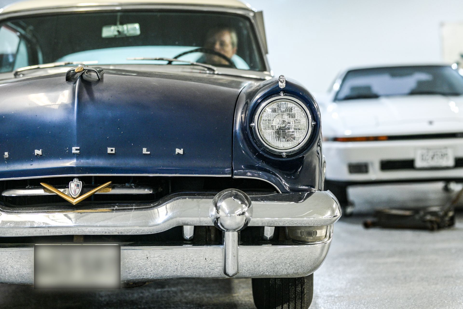 Dark blue classic Lincoln car in a garage, with a white sports car in the background. A person is in the driver's seat.