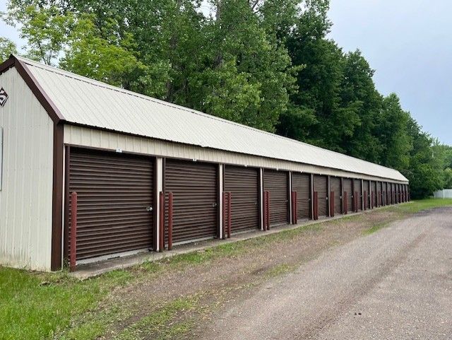 Row of brown storage units with metal roll-up doors, white walls, and a brown roof. They are alongside a gravel road and wooded area.