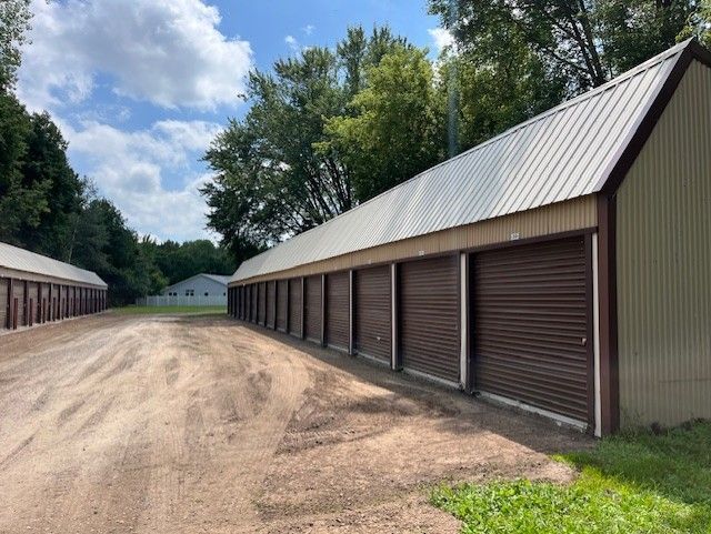 Storage units in a row with brown doors and a light-colored metal roof, set on a dirt and gravel lot. Trees in the background.