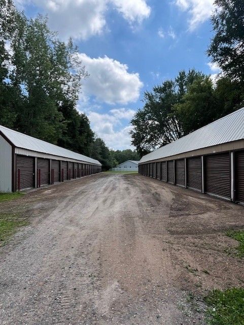 Gravel drive between two rows of storage units with brown doors and white metal roofs under a partly cloudy sky.