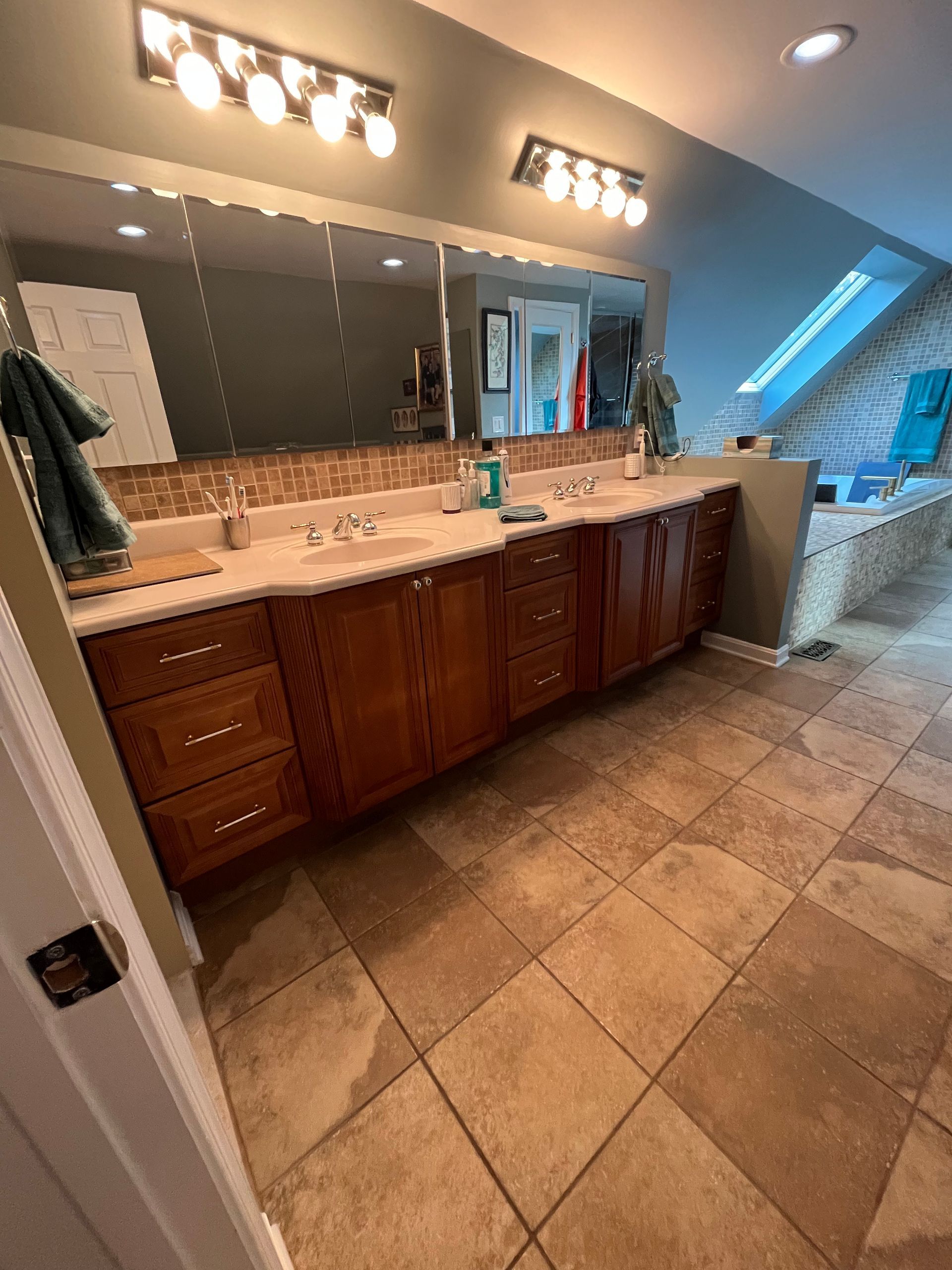 Bathroom with wood cabinets, light countertops, large mirrors, and beige tiled floor.