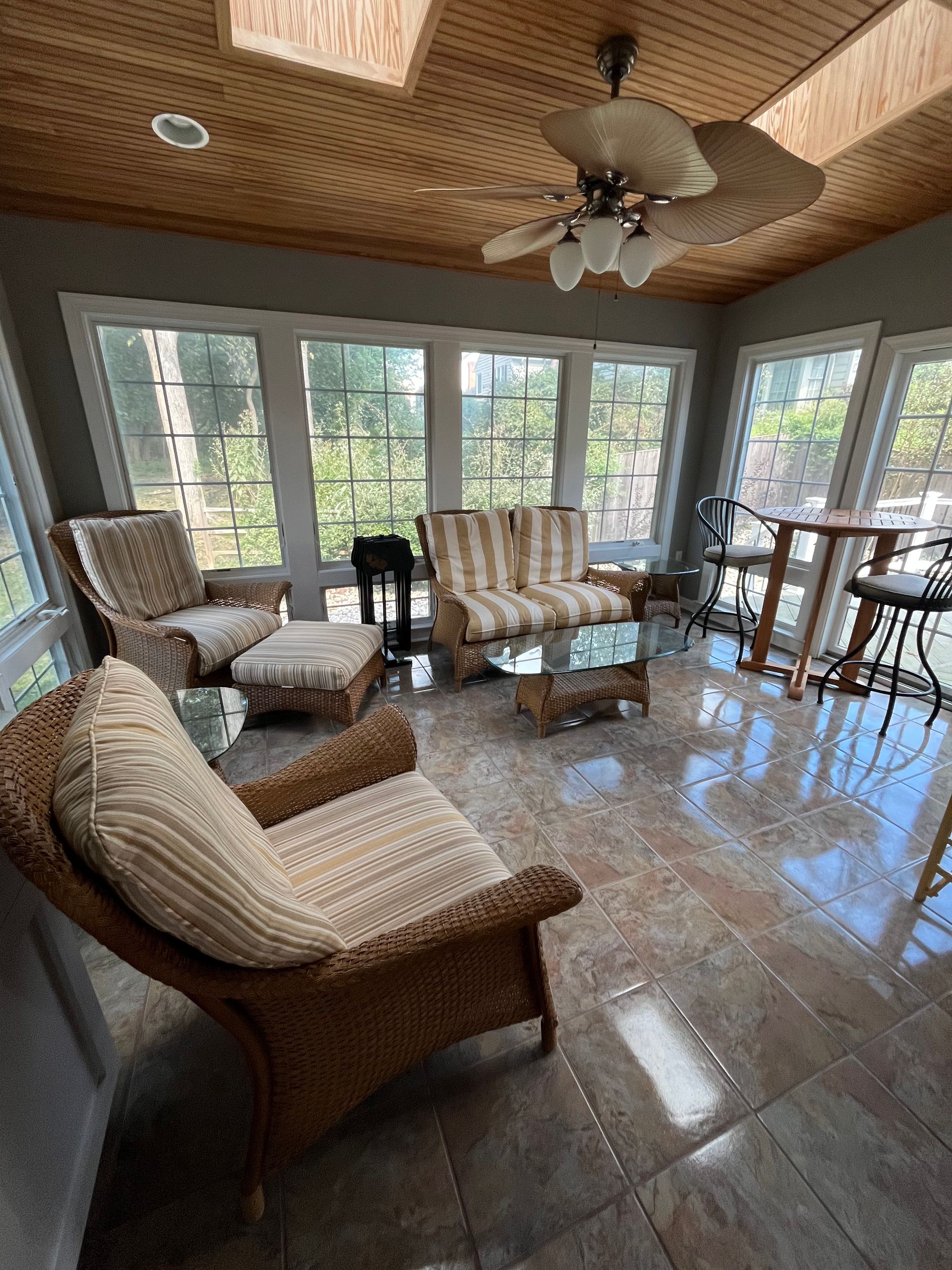 Sunroom with wicker furniture, glass-topped coffee table, windows, wooden ceiling, and tile floor.