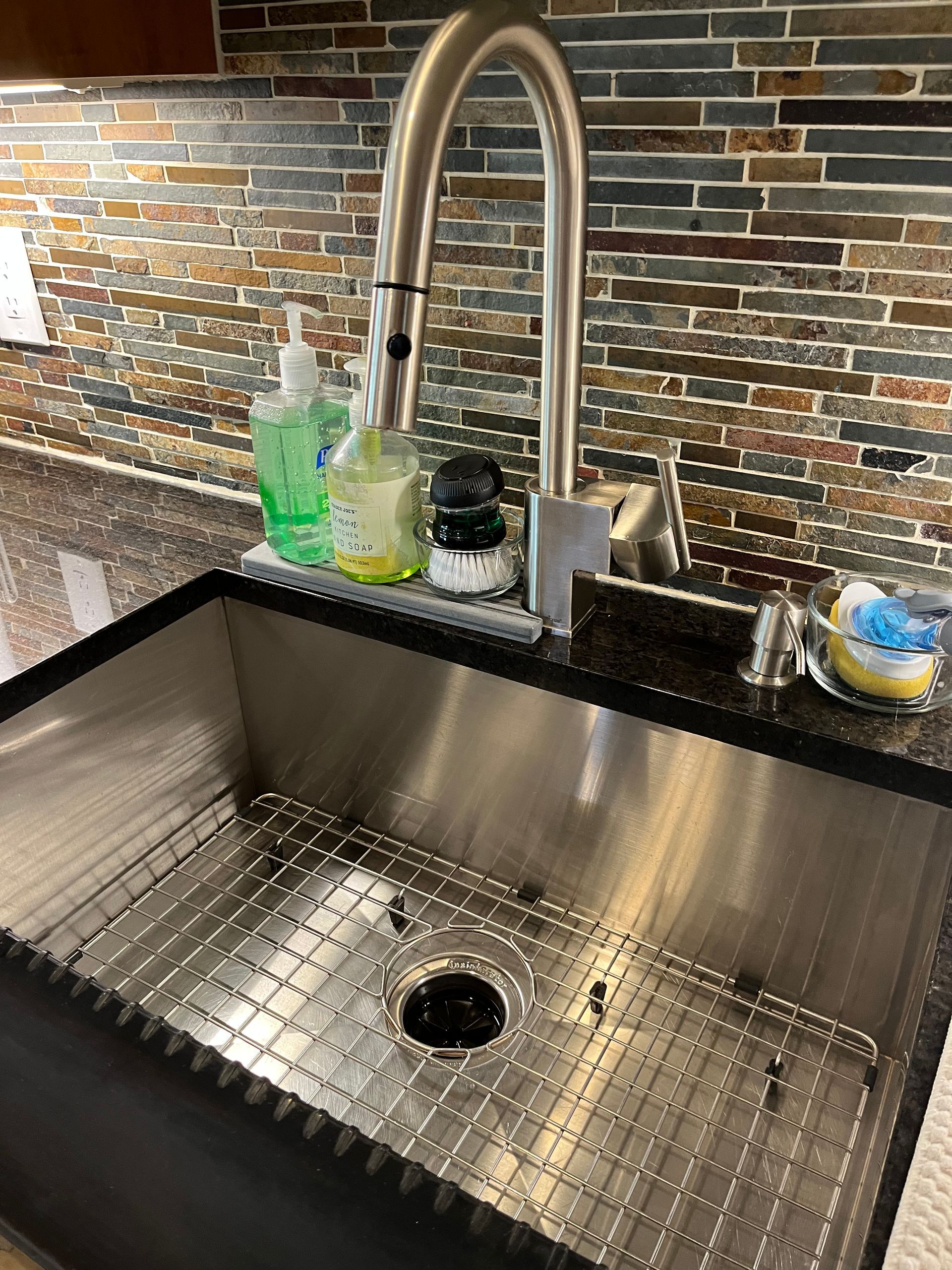 Stainless steel kitchen sink with grid, faucet, and soap dispensers against a mosaic tile backsplash.