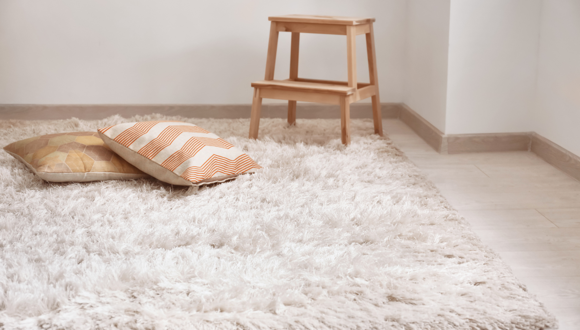 A room with a white rug , pillows and a wooden stool.