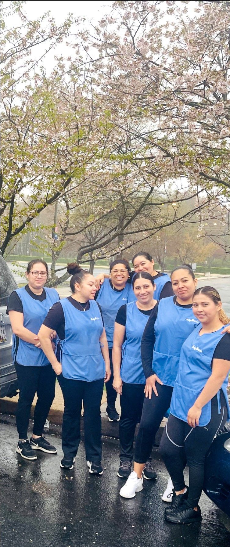 A group of women in blue vests and black pants pose under a tree with pink blossoms.