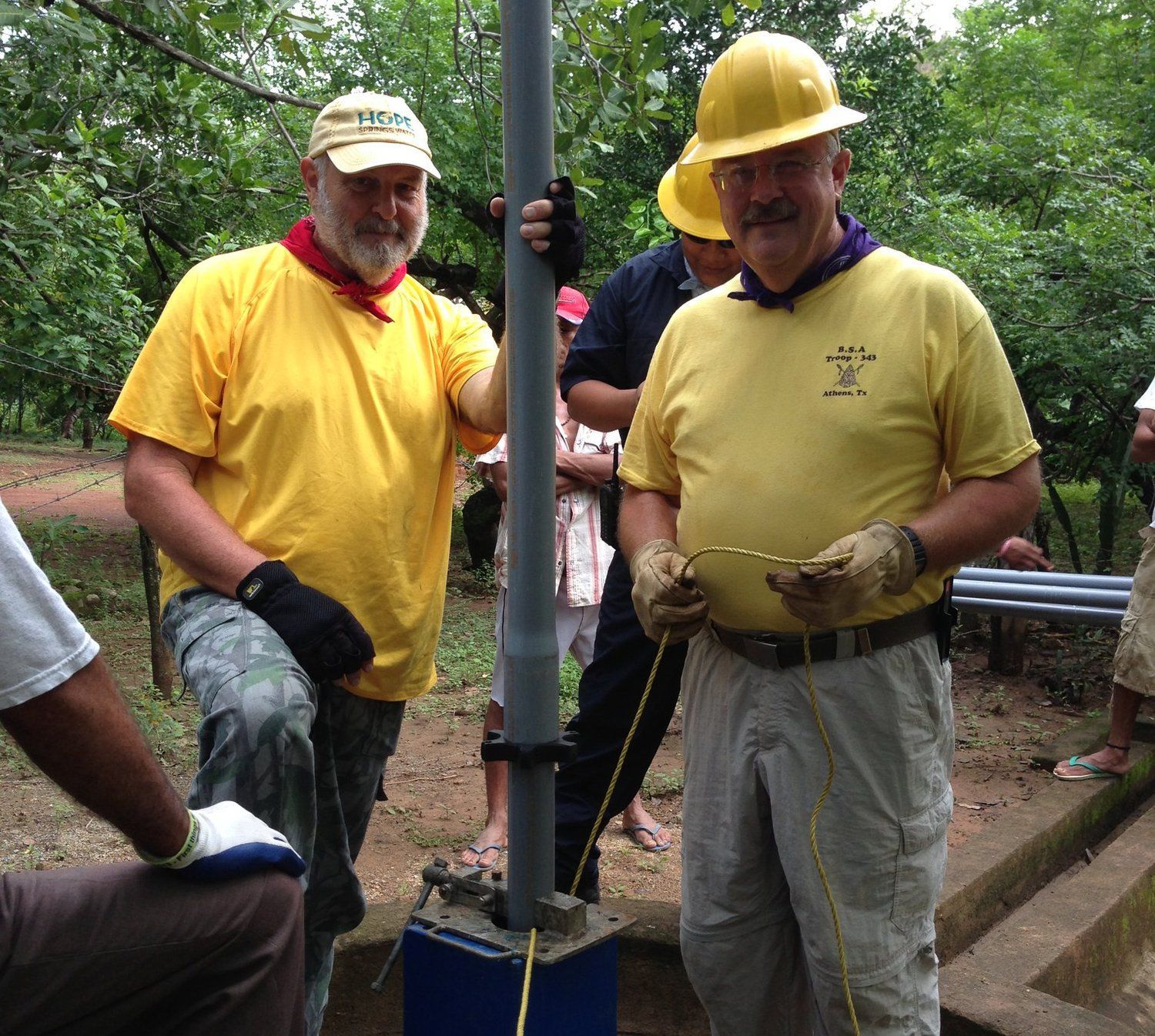 Two men wearing yellow shirts and hard hats are working on a pole