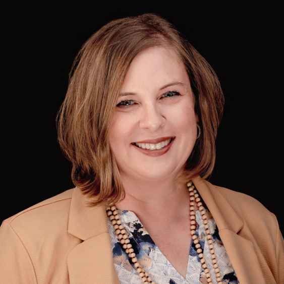 A woman wearing a tan jacket and a necklace is smiling for the camera.