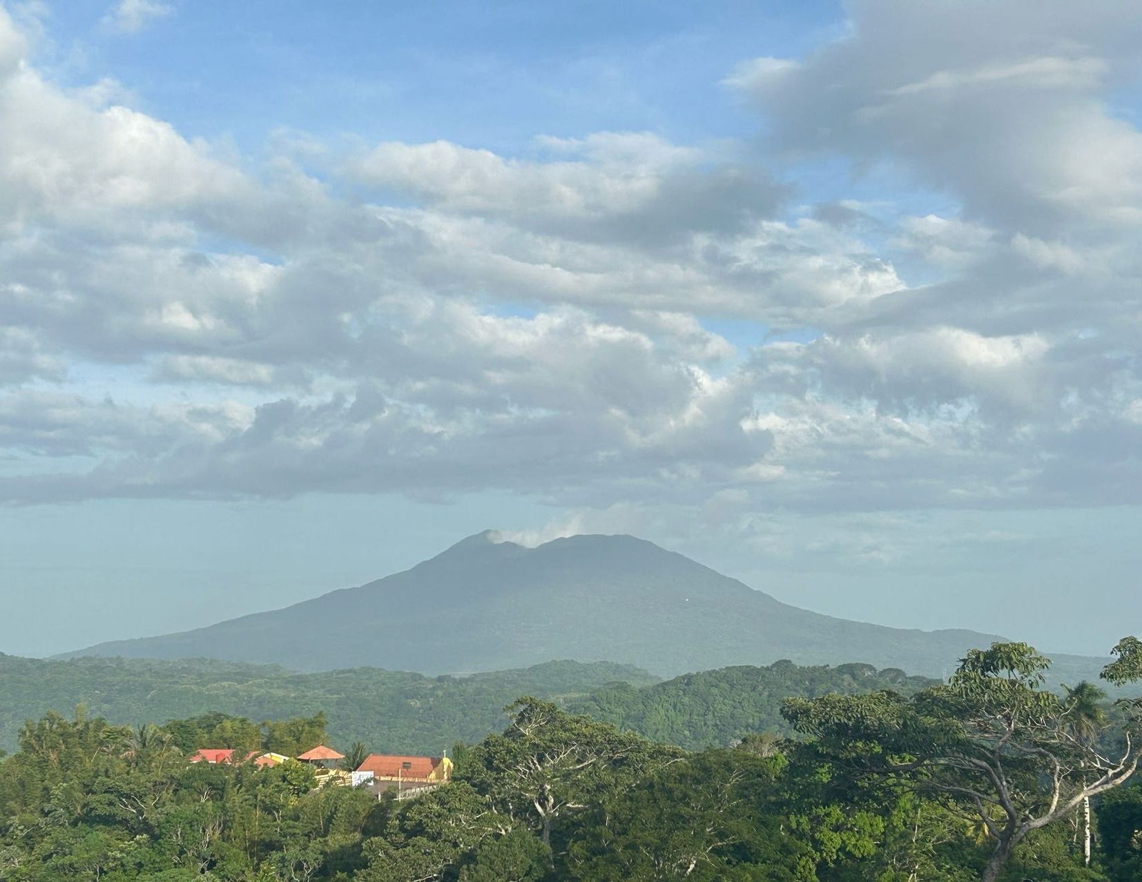 A green forest with a volcano in the background and a blue sky with clouds