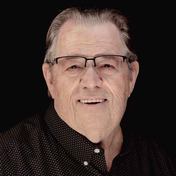 An elderly man wearing glasses and a black shirt is smiling for the camera.