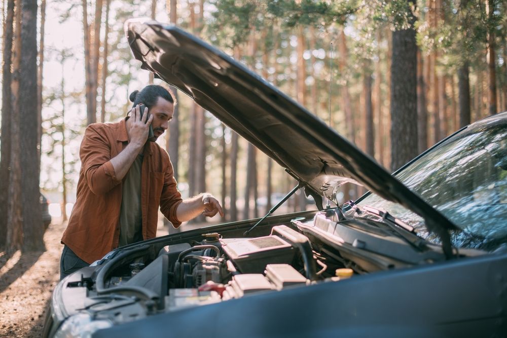 A man is talking on a cell phone while looking under the hood of his broken down car.