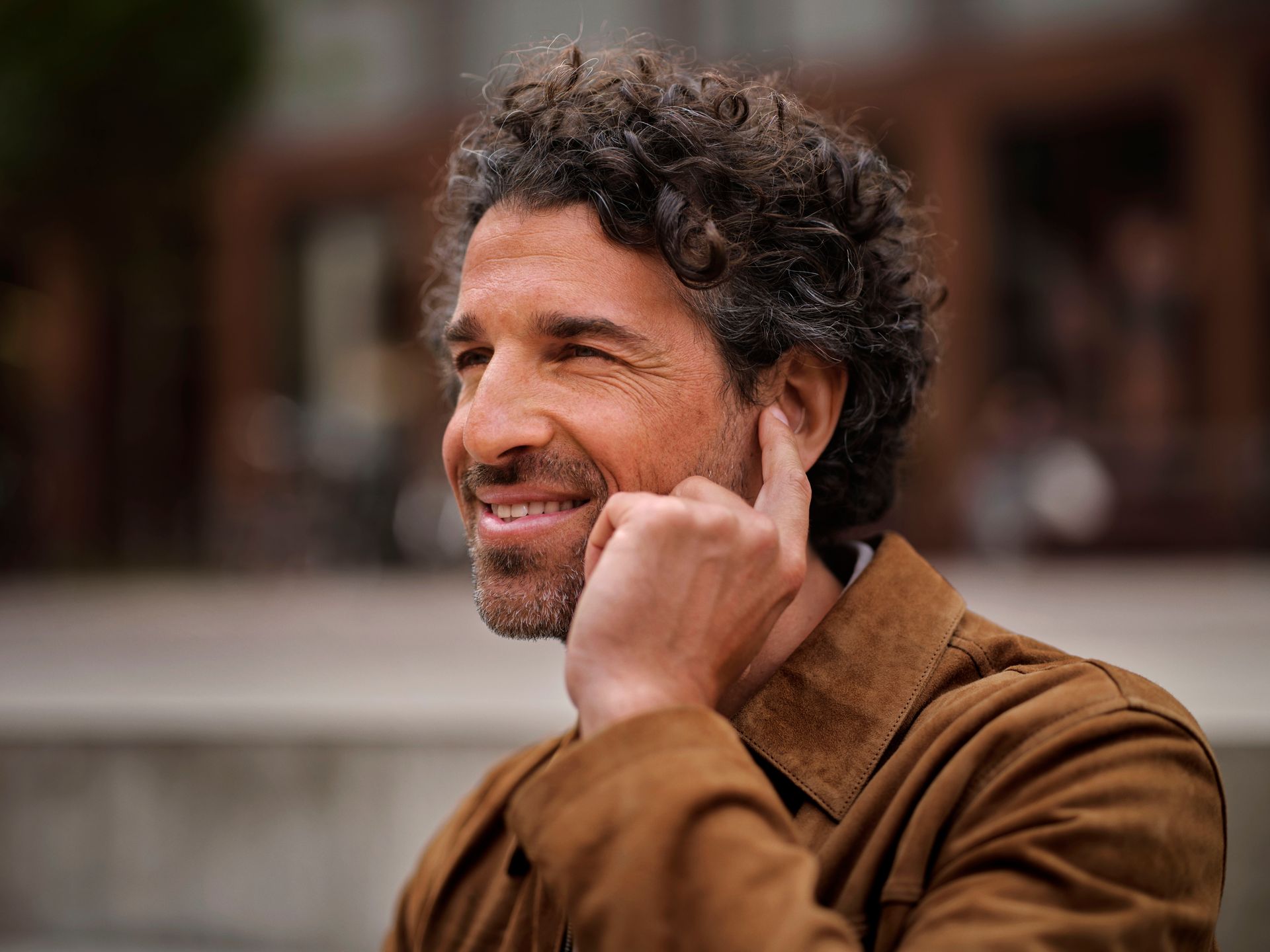 Maður með krullað hár í brúnum jakka, brosandi og snertir eyrað á sér, úti.