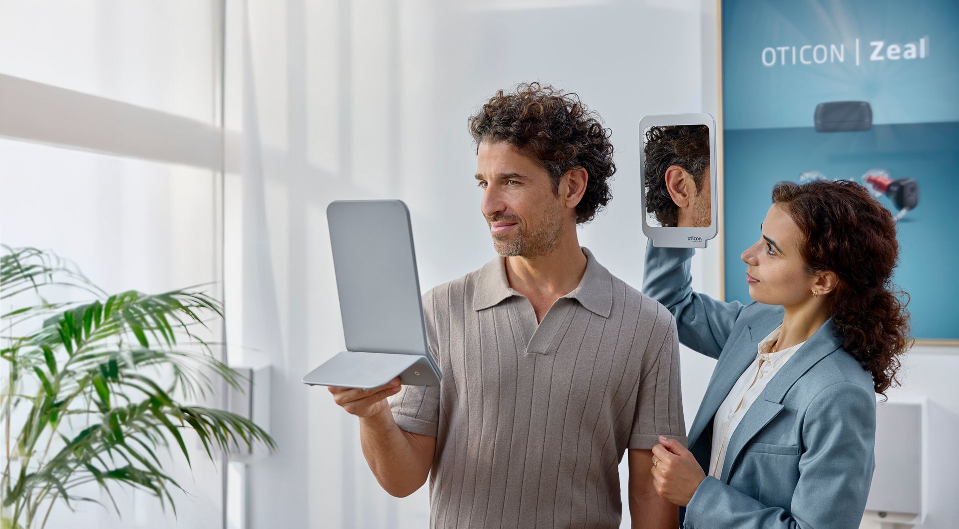 Maður heldur á fartölvu, kona heldur á spegli, innandyra við glugga með grænu umhverfi og auglýsingu fyrir heyrnartæki.