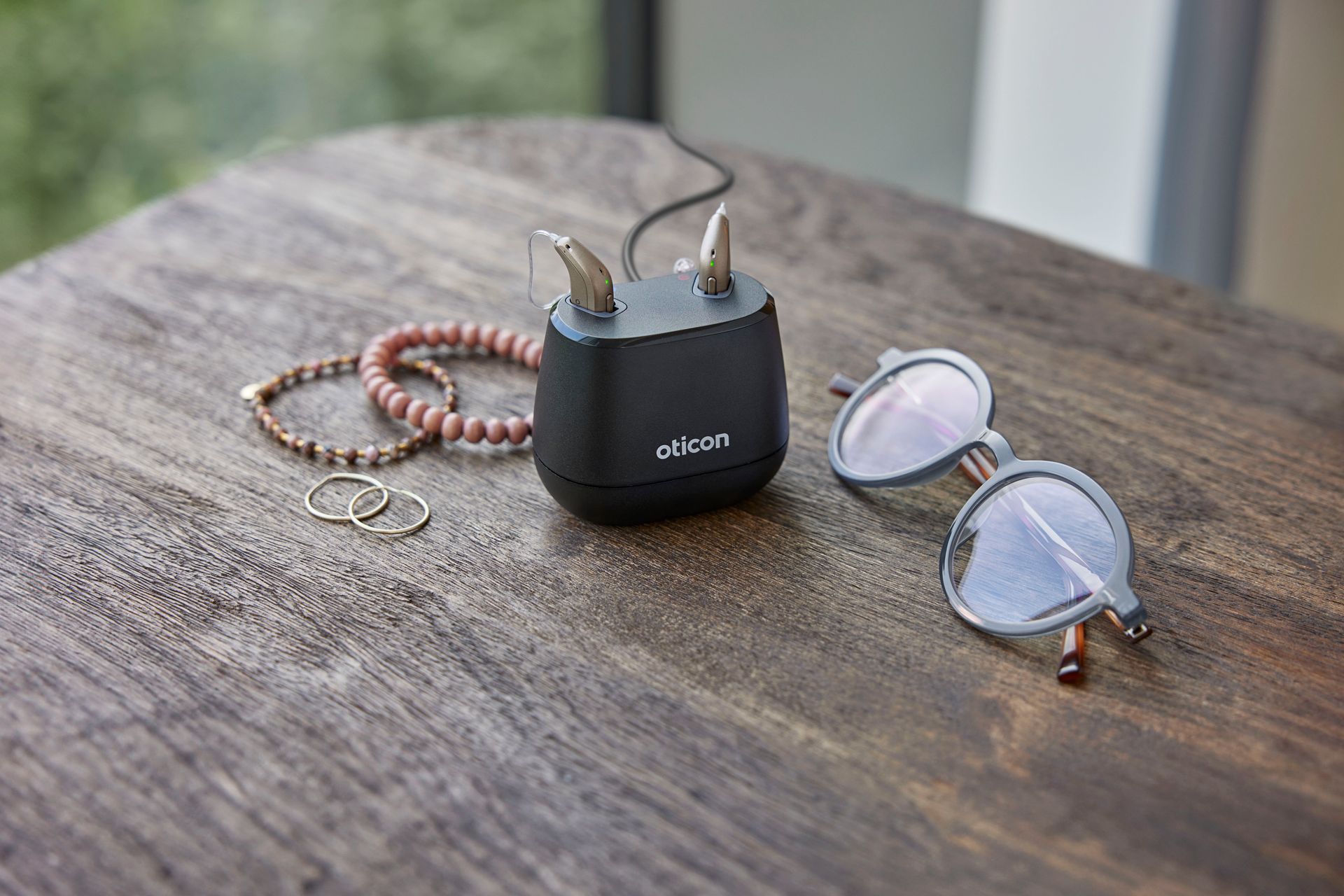 Svart hleðslutæki fyrir heyrnartækja með heyrnartækjum, gleraugum og skartgripum á viðarfleti.