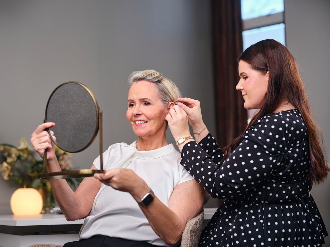 Kona horfir í spegil á meðan önnur kona lagar hárið á sér. Innandyra, hlutlaust umhverfi.