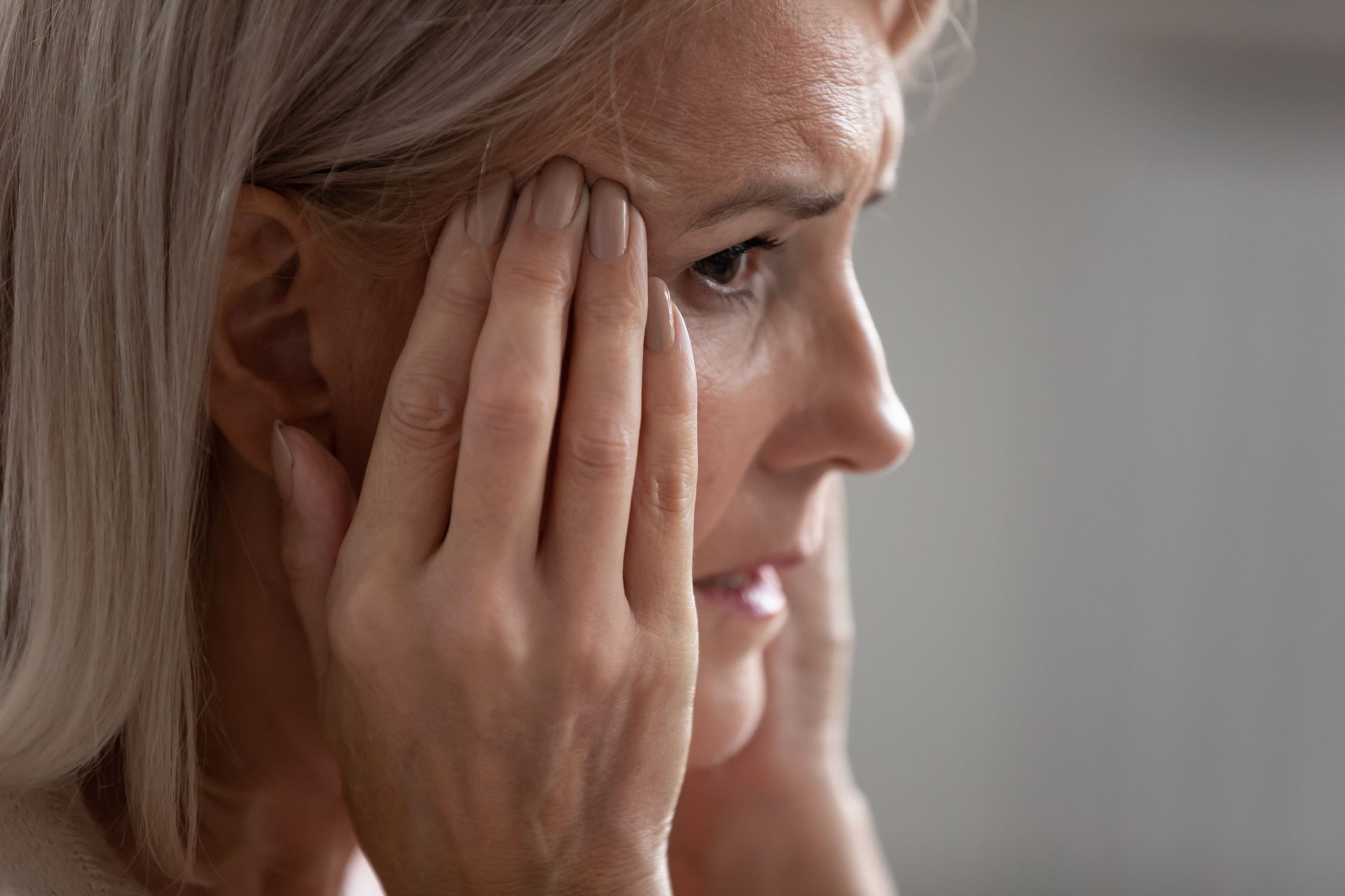Kona með grátt hár, hendur á gagnaugum, andlit sýnir sársauka og áhyggjur.