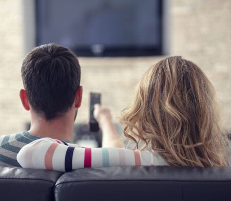 Par situr í sófa og horfir á sjónvarp; kona heldur á fjarstýringu.