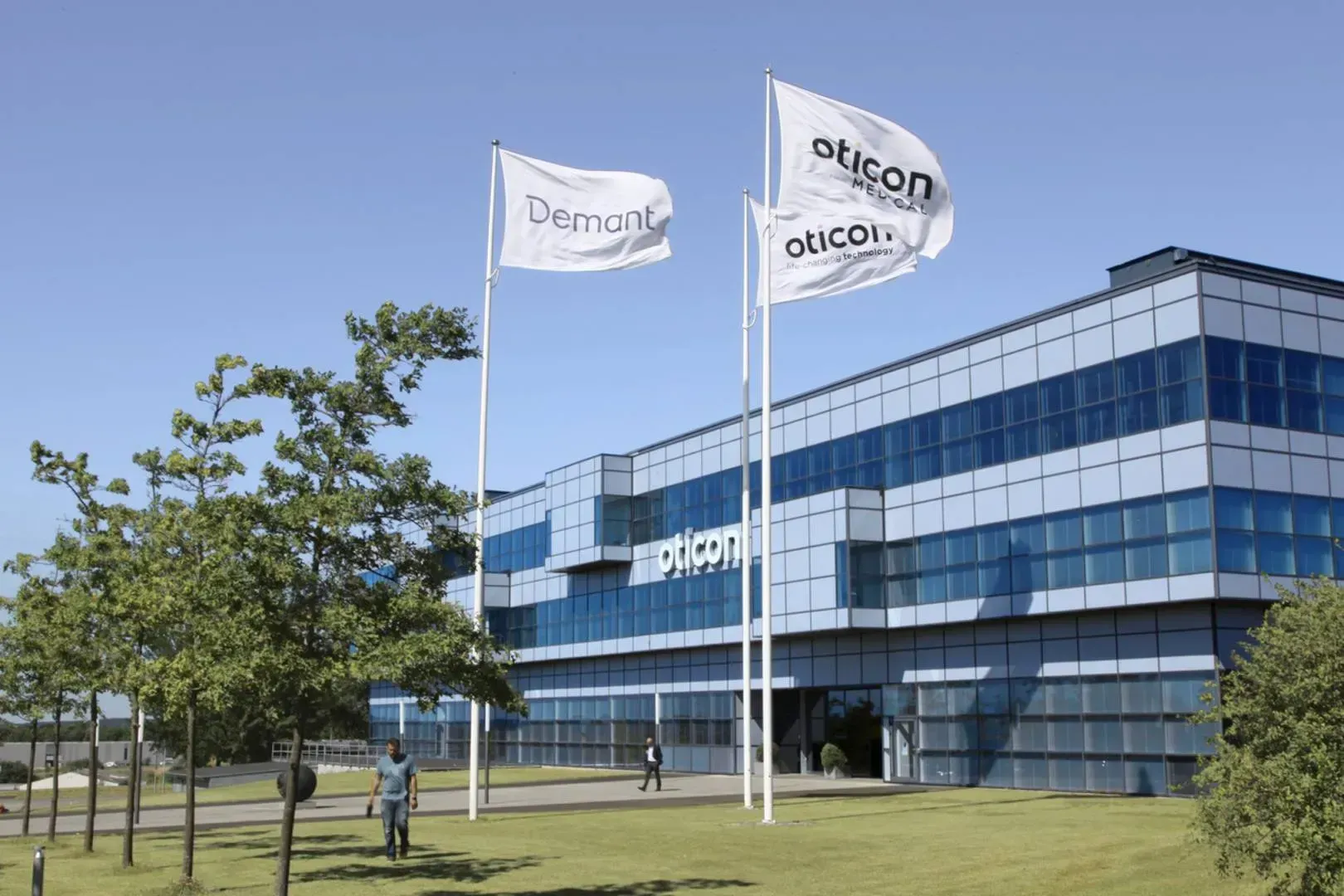 Nútímaleg bygging með fánum með merkjunum „Demant“ og „Oticon“. Tveir ganga um lóðina.