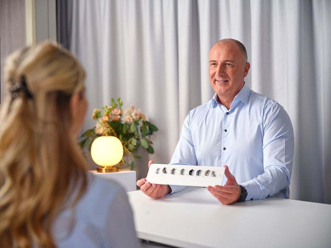 Maður með heyrnartæki í höndunum, brosandi til konu, innandyra með blómum og gluggatjöldum.