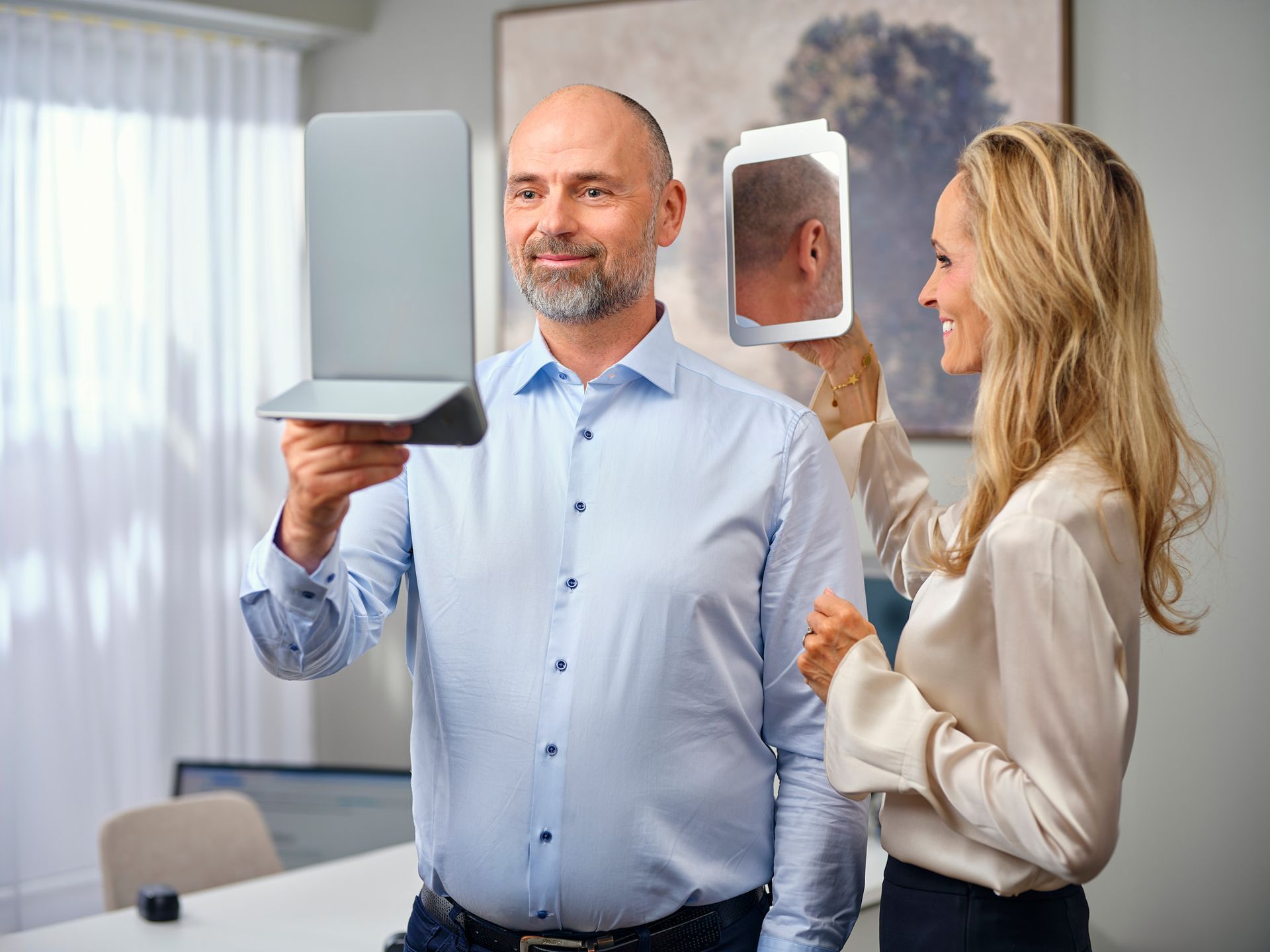 Maður heldur á fartölvu, kona heldur á spegli, innandyra við glugga með grænu umhverfi og auglýsingu fyrir heyrnartæki.