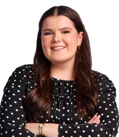 Kona með sítt brúnt hár, brosandi, í svartri doppóttri blússu, með krosslagða hendur.