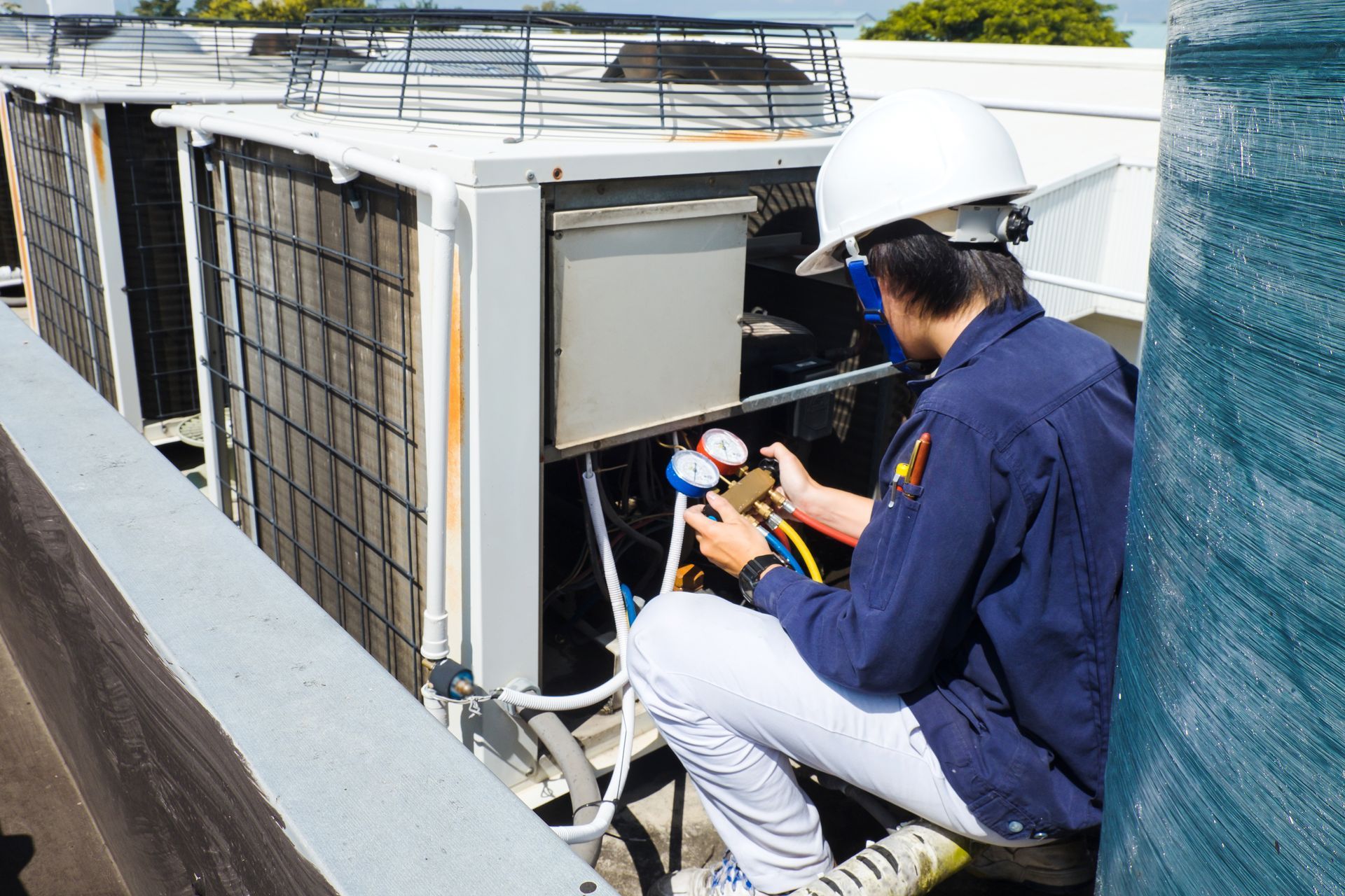 Technician in a hard hat kneeling on a rooftop, using a manifold gauge set to service an HVAC condenser unit. Technician in a hard hat kneeling on a rooftop, using a manifold gauge set to service an HVAC condenser unit.