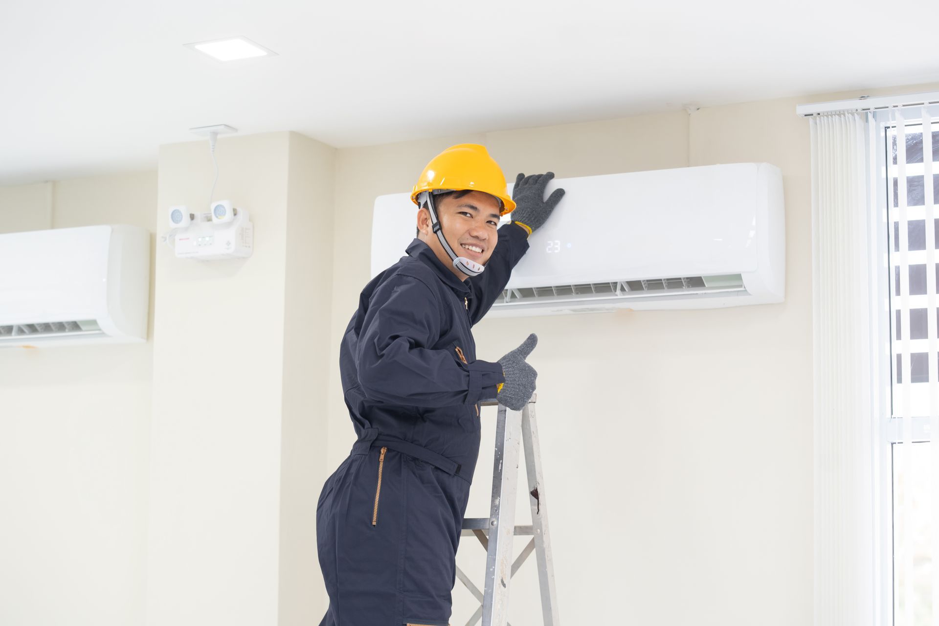 A technician in a hard hat and uniform on a ladder, smiling and giving a thumbs-up while working on a wall-mounted AC unit. A technician in a hard hat and uniform on a ladder, smiling and giving a thumbs-up while working on a wall-mounted AC unit.