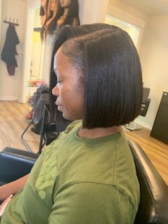 Woman with a side-parted, sleek, black bob hairstyle. She wears a green shirt, sitting in a salon chair.