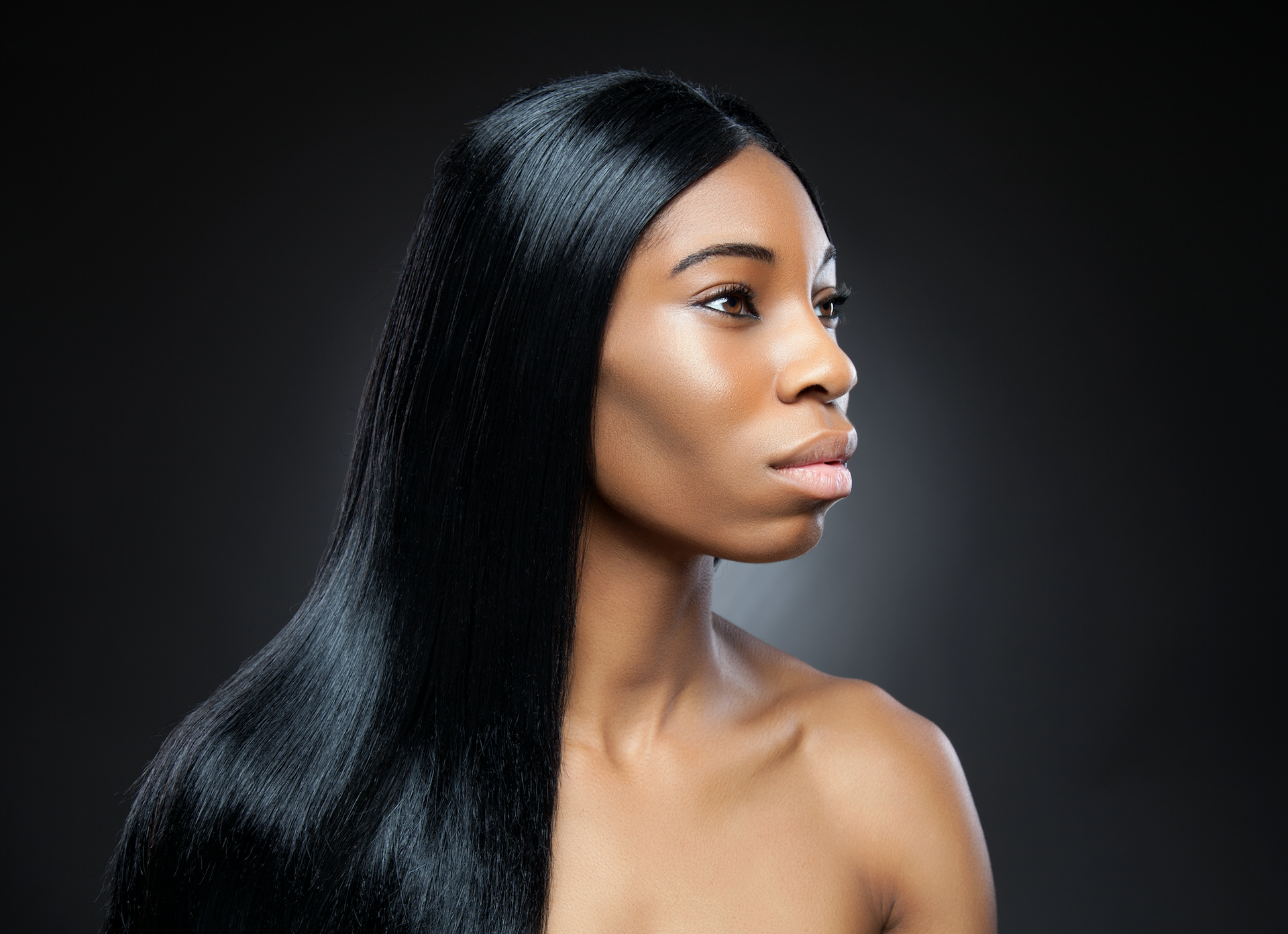 Woman with long, sleek black hair looking right, bare shoulders, against a dark gray background.