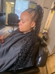 Person with braided hair and curly extensions, wearing a black cape, in a salon chair.
