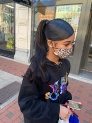 Woman in a black hoodie and patterned mask, hair in a high ponytail, holding a phone, walking outside.