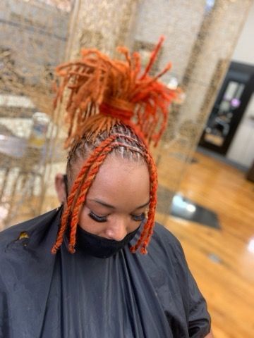 Person with orange-dyed locs styled in a top bun and face-framing braids, wearing a black face mask and salon cape.