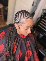 Woman with cornrows styled in an intricate pattern, braids flowing down. Wearing a red and black floral cape, indoors.