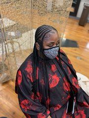 Person with braided hair and a patterned face mask, wearing a red-floral robe in a salon setting.