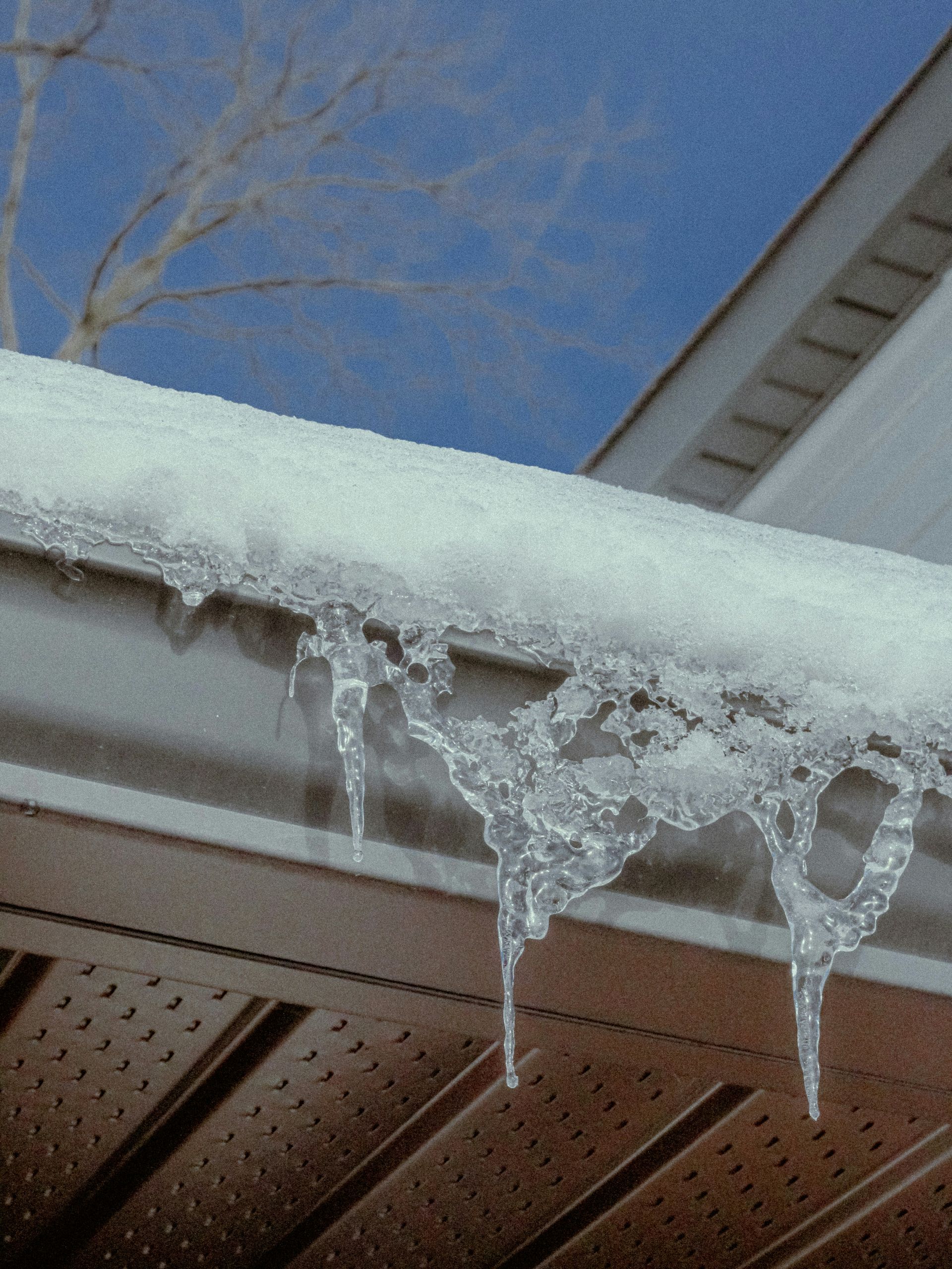 Ice and icicles hang from a brown gutter on a roof against a blue sky.