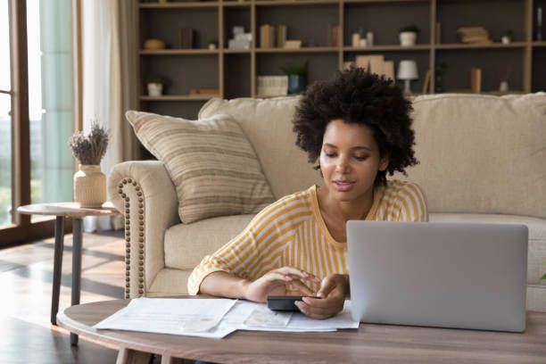Woman makes calculations while sitting in a low table next to her laptop, in a living room.