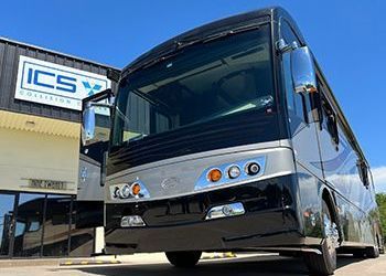 A large black and silver bus is parked in front of a building that says ics.