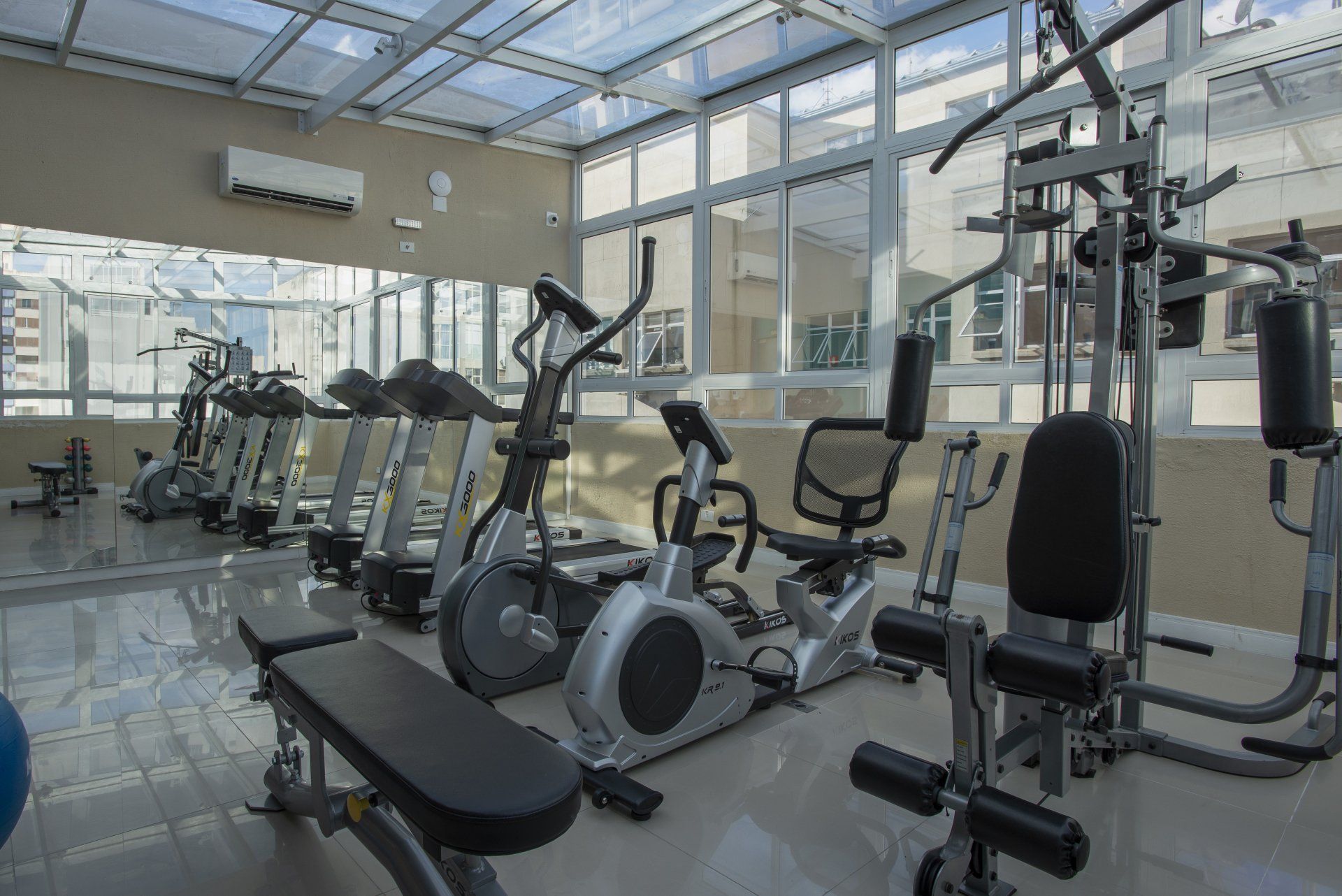 Academia com esteiras, elípticos, aparelhos de musculação e um banco, sob um teto de vidro.