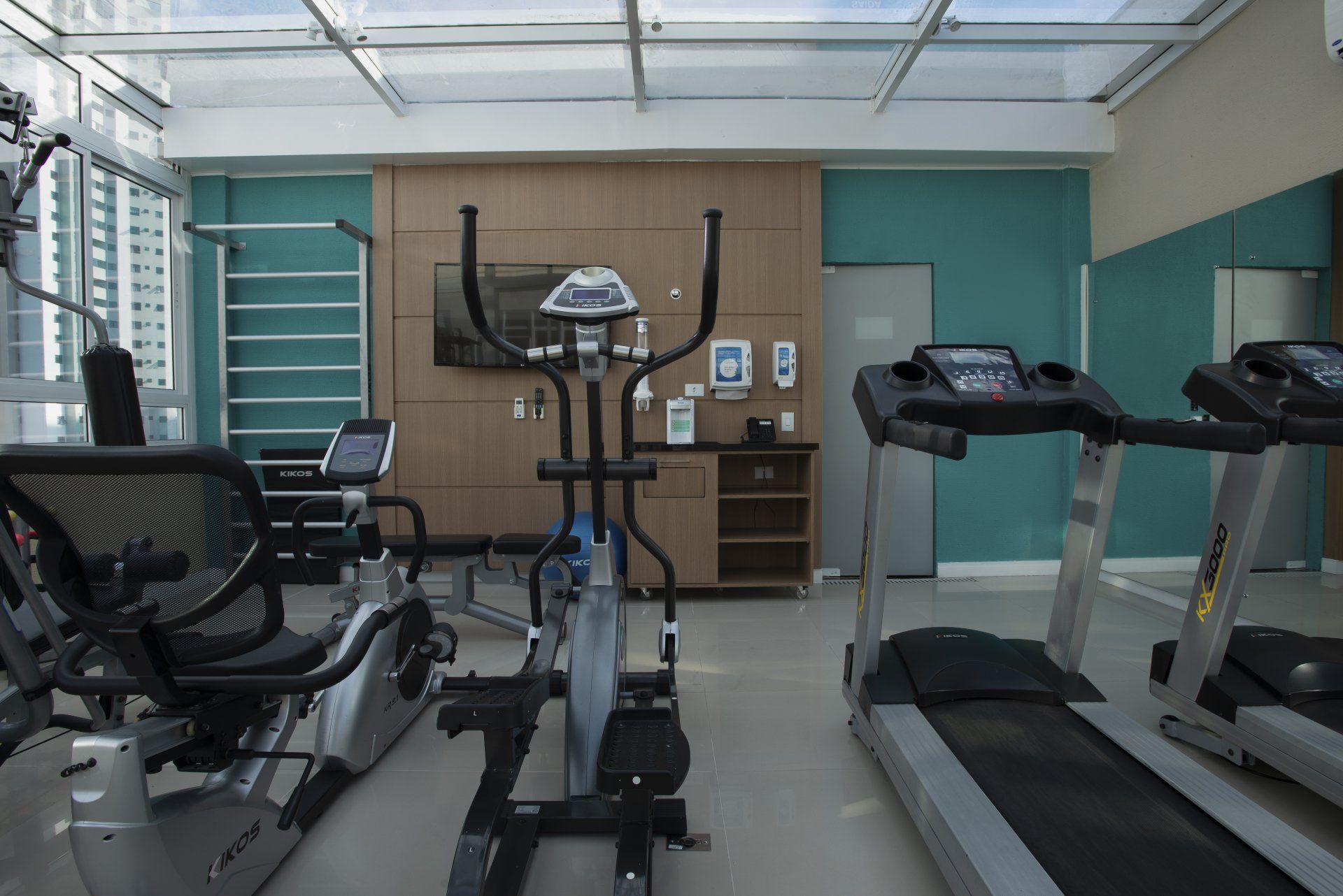 Academia com esteiras, elíptico, bicicleta ergométrica, TV e barras de exercícios. Paredes em tons turquesa e painéis de madeira.