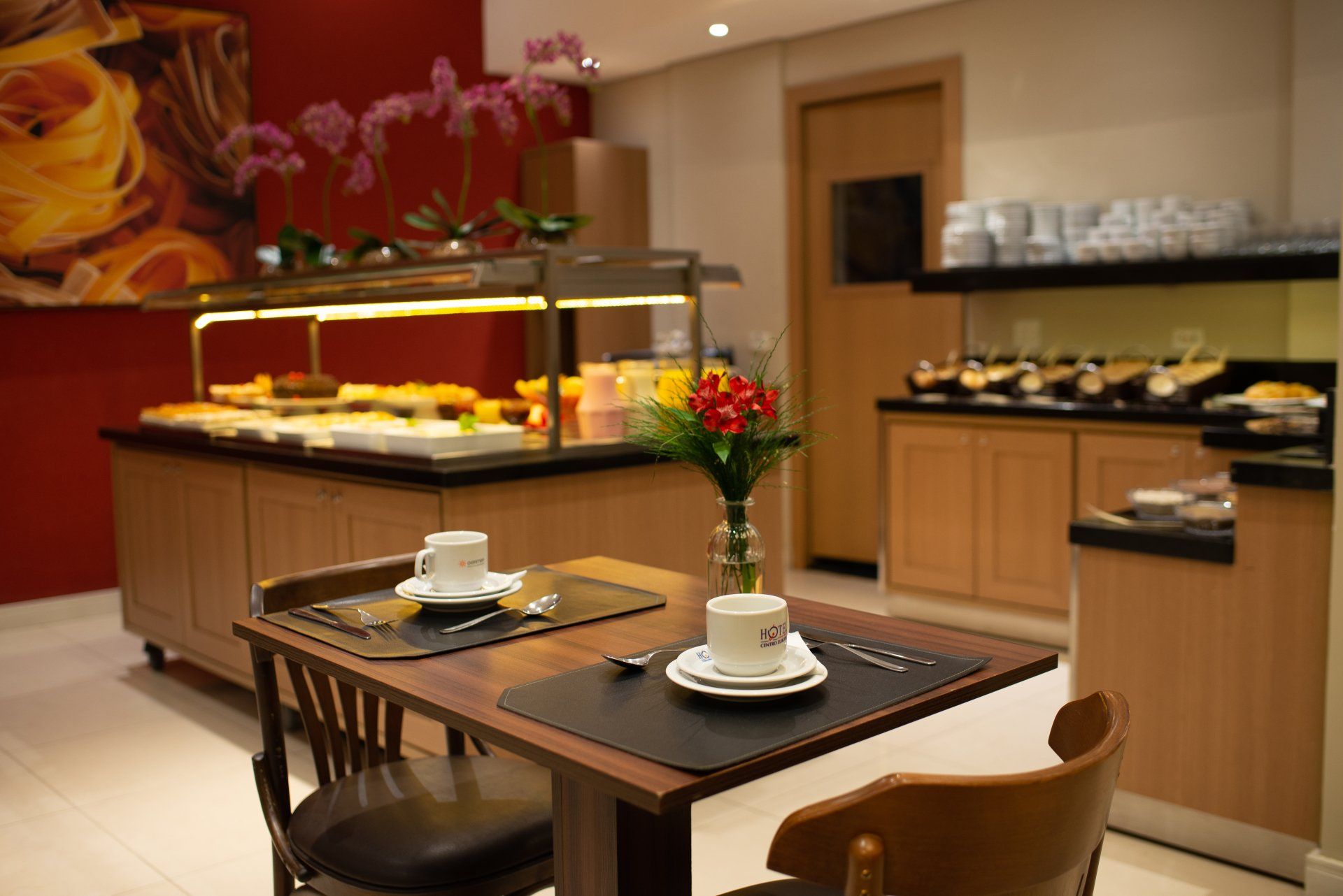 Uma área de café da manhã do hotel com doces, uma mesa posta para dois e um buquê de flores.