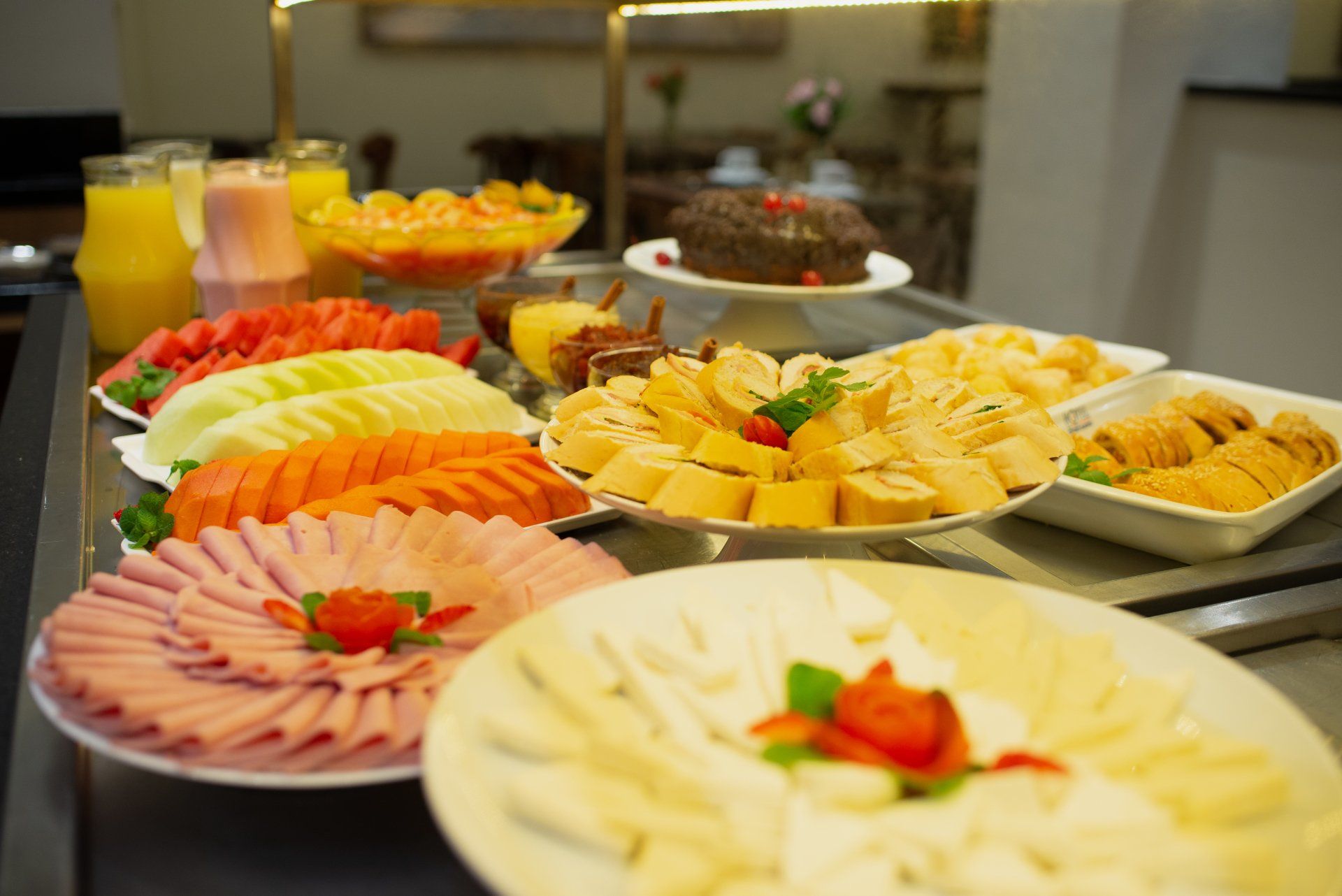 Mesa de bufê com alimentos variados, incluindo frutas fatiadas, presunto, queijo, doces e bebidas.