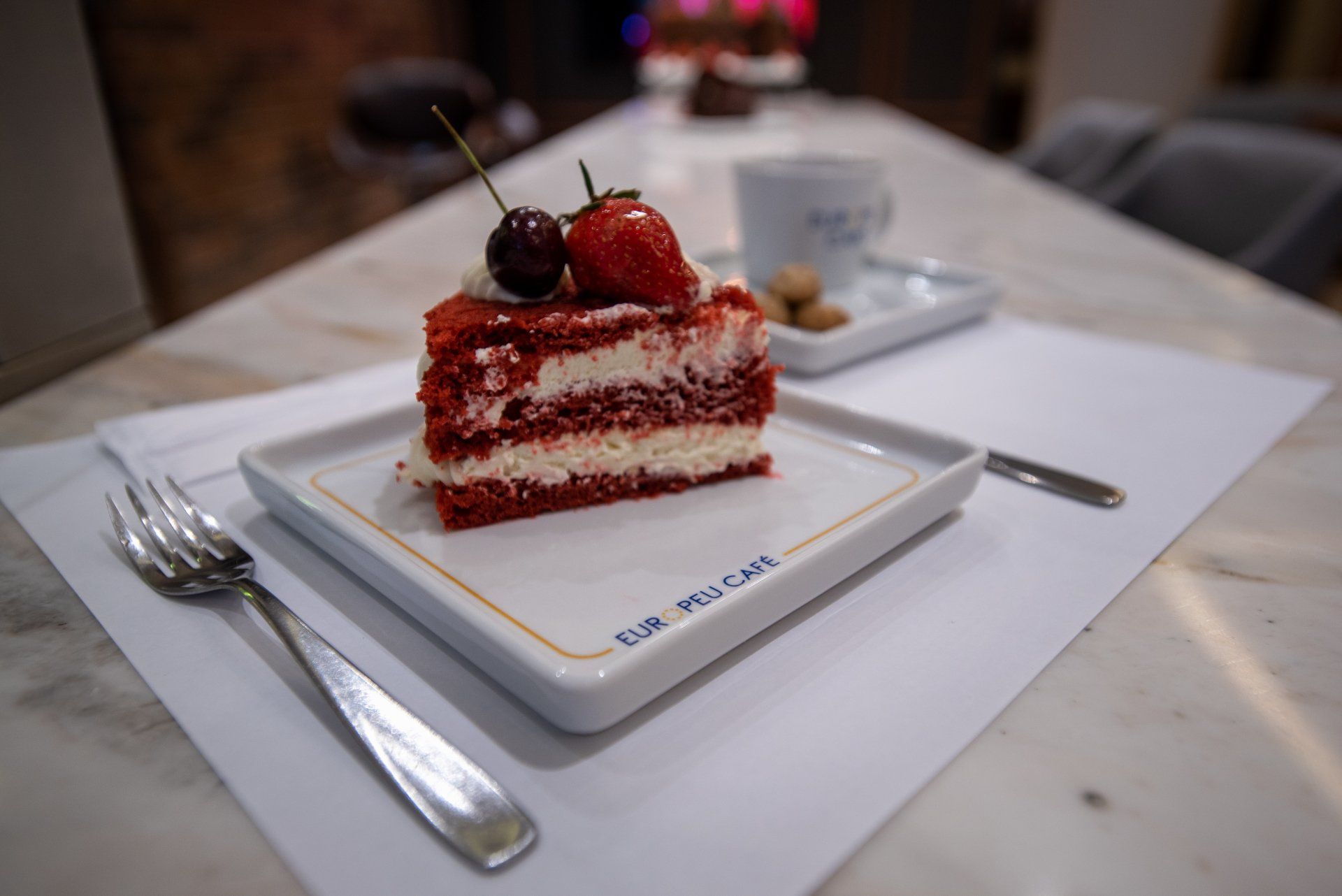 Fatia de bolo red velvet em um prato branco, coberto com cerejas e morangos. Ao lado de um garfo sobre uma mesa de mármore.