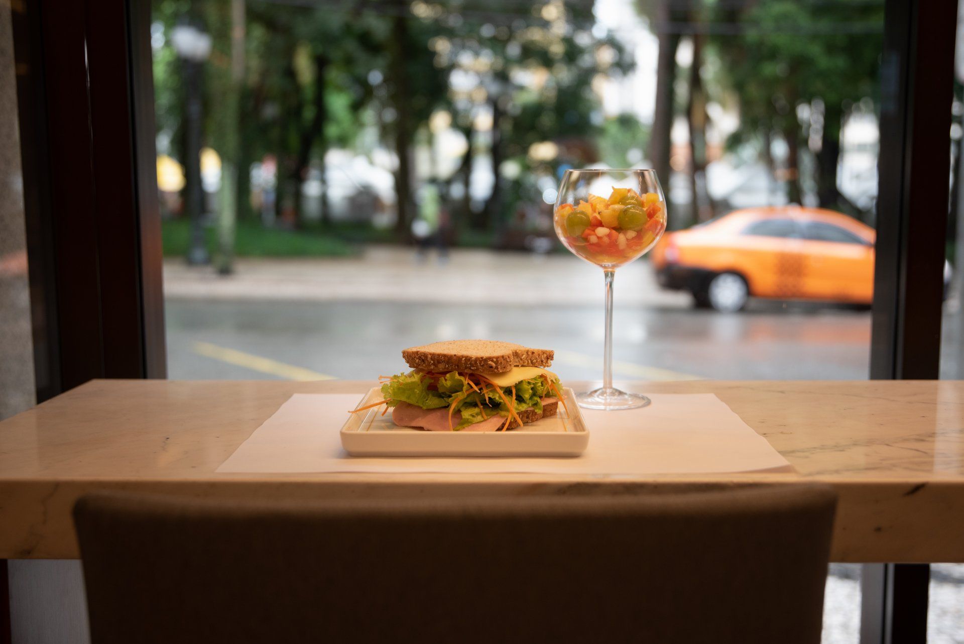 Sanduíche e salada de frutas em uma mesa em frente a uma janela com vista para uma rua com um táxi amarelo.
