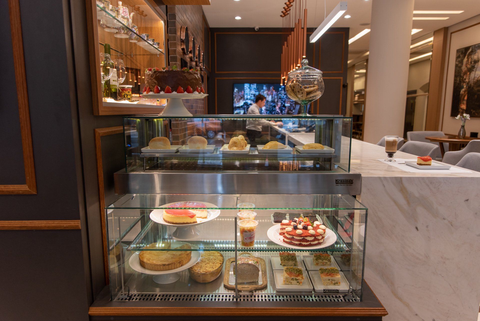 Vitrine de doces em um café, exibindo diversas sobremesas. O café tem decoração moderna e iluminação suave.
